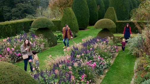 Visitors walking between borders of pink and purple flowers and yew topiary, in autumn at Hinton Ampner
